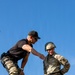 Two individuals are positioned at the top of a wooden structure, likely a rappelling or obstacle course tower. An instructor in a black t-shirt and camouflage pants is leaning toward a U.S. Army soldier wearing a helmet marked '517' and full OCP (Operational Camouflage Pattern) uniform with rappelling harness and rope. The interaction appears to be instructional guidance prior to a rappel descent.
