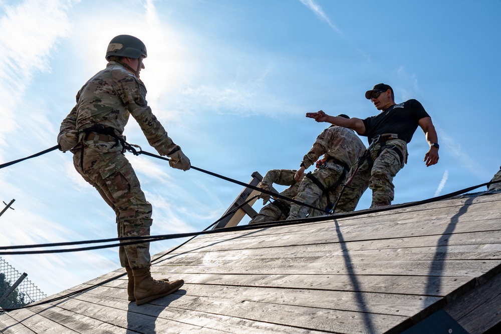 Military personnel are engaged in a rappelling or rope descent training exercise on a wooden inclined structure. An instructor in civilian black clothing and cap is directing a soldier in full OCP camouflage uniform and helmet who is managing ropes on the sloped surface. At least three individuals are visible, with safety harnesses and ropes in use throughout the exercise.