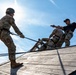 Military personnel are engaged in a rappelling or rope descent training exercise on a wooden inclined structure. An instructor in civilian black clothing and cap is directing a soldier in full OCP camouflage uniform and helmet who is managing ropes on the sloped surface. At least three individuals are visible, with safety harnesses and ropes in use throughout the exercise.