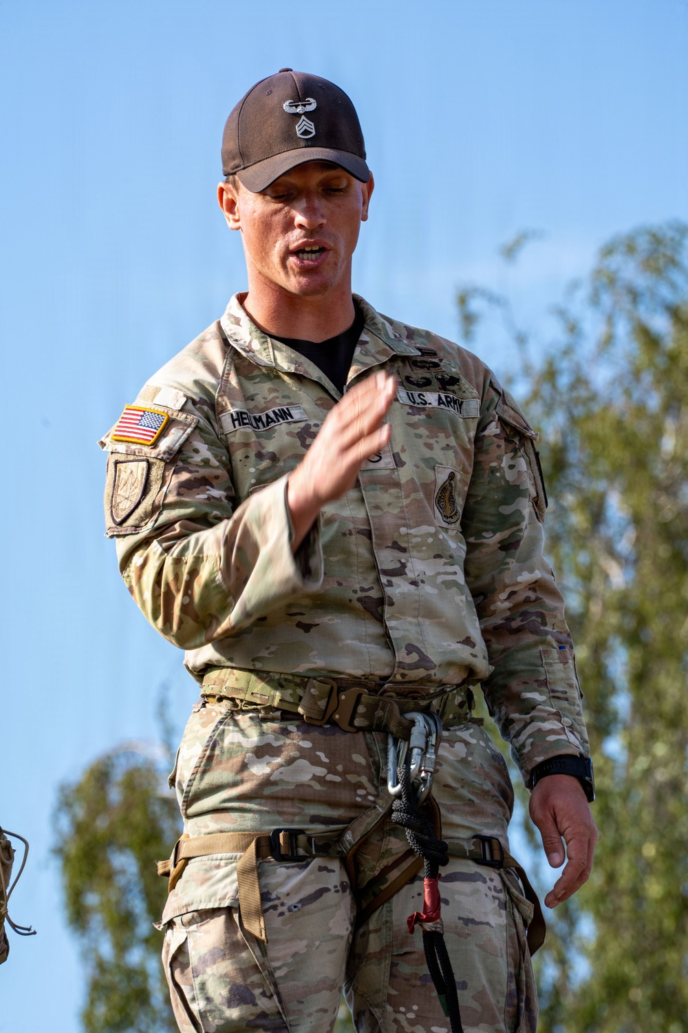 A U.S. Army soldier identified by name tape as 'Heilmann' is speaking or giving instruction while wearing OCP (Operational Camouflage Pattern) uniform. He is equipped with a rappelling or climbing harness with carabiners attached, suggesting a training or demonstration context. He wears a dark baseball cap with what appears to be a Staff Sergeant rank insignia and a Combat Action Badge.