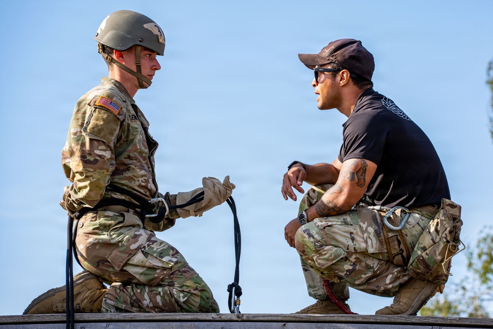 Two individuals are engaged in what appears to be a rappelling or rope training exercise at an elevated position. One person is wearing full military uniform with a helmet and harness, while the other, dressed in civilian-style tactical clothing with a baseball cap and sunglasses, appears to be an instructor providing guidance. They are interacting face-to-face, with a rope or cord visible between them.