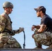 Two individuals are engaged in what appears to be a rappelling or rope training exercise at an elevated position. One person is wearing full military uniform with a helmet and harness, while the other, dressed in civilian-style tactical clothing with a baseball cap and sunglasses, appears to be an instructor providing guidance. They are interacting face-to-face, with a rope or cord visible between them.