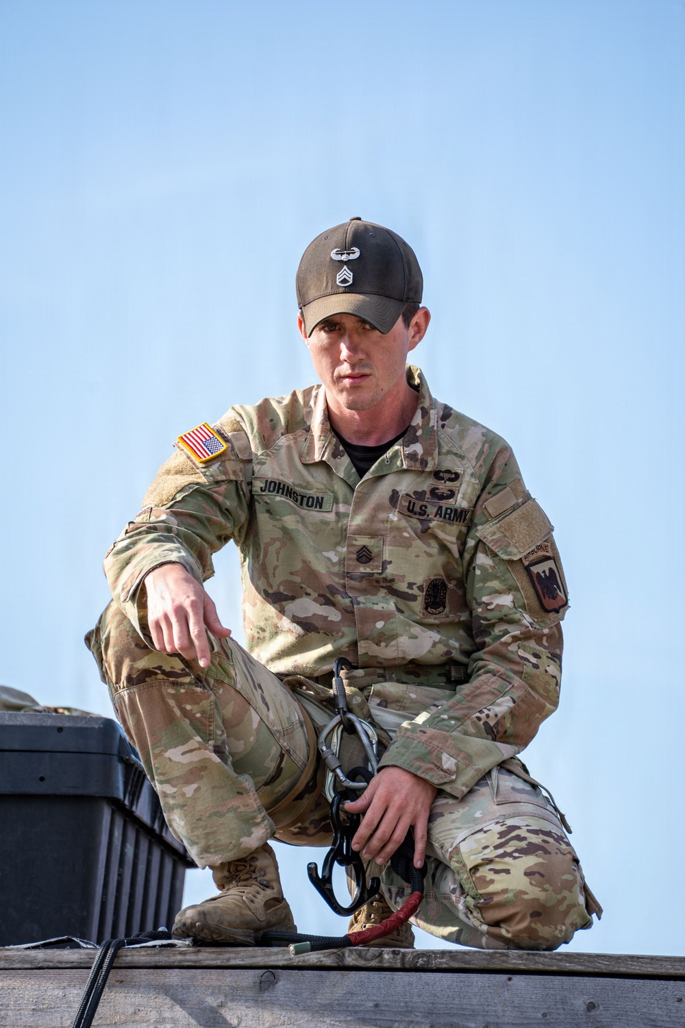 A U.S. Army soldier identified by name tape as 'Johnston' is crouching on an elevated wooden platform, holding carabiners and rigging equipment. He is wearing OCP (Operational Camouflage Pattern) uniform with visible rank insignia of Staff Sergeant, an Airborne tab, U.S. flag patch, and a Combat Action Badge on his patrol cap. The image appears to be taken during a training or operational activity involving rappelling or rigging.