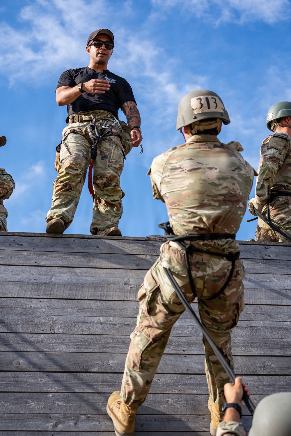 A military training exercise is underway at what appears to be a rappelling or obstacle course wall. An instructor in a black t-shirt and camouflage pants stands atop a wooden wall, gesturing instructions to trainees below. At least two trainees wearing helmets and harnesses are visible, with one numbered '313' actively climbing or rappelling the wooden structure.