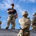 A military training exercise is underway at what appears to be a rappelling or obstacle course wall. An instructor in a black t-shirt and camouflage pants stands atop a wooden wall, gesturing instructions to trainees below. At least two trainees wearing helmets and harnesses are visible, with one numbered '313' actively climbing or rappelling the wooden structure.