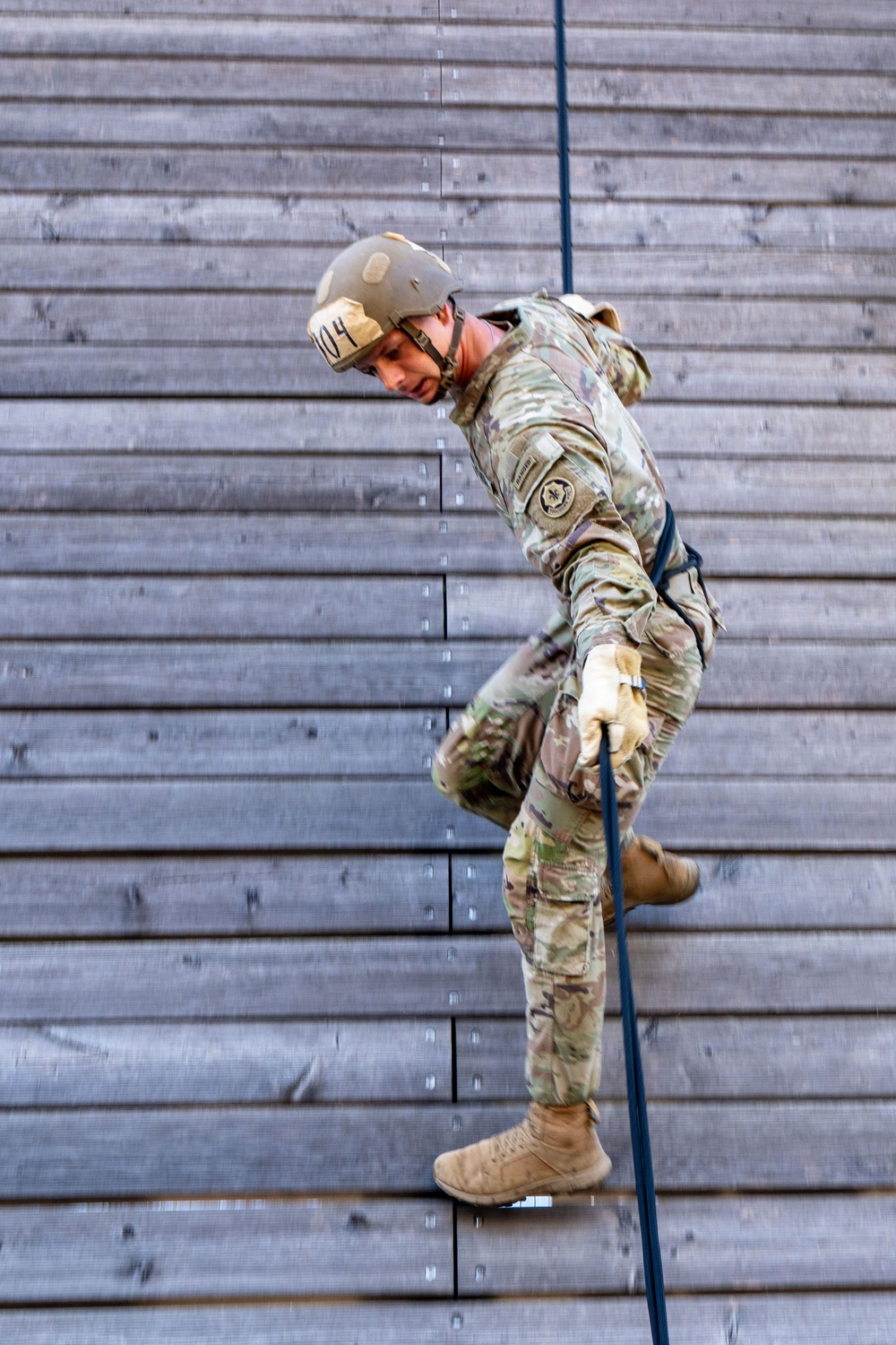 A military service member in full camouflage uniform is rappelling down a large wooden wall structure. The individual is wearing a helmet marked with the number '04' and is gripping a blue rope while descending in a controlled posture. This appears to be a training exercise at a military obstacle or rappel tower.