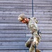 A military service member in full camouflage uniform is rappelling down a large wooden wall structure. The individual is wearing a helmet marked with the number '04' and is gripping a blue rope while descending in a controlled posture. This appears to be a training exercise at a military obstacle or rappel tower.