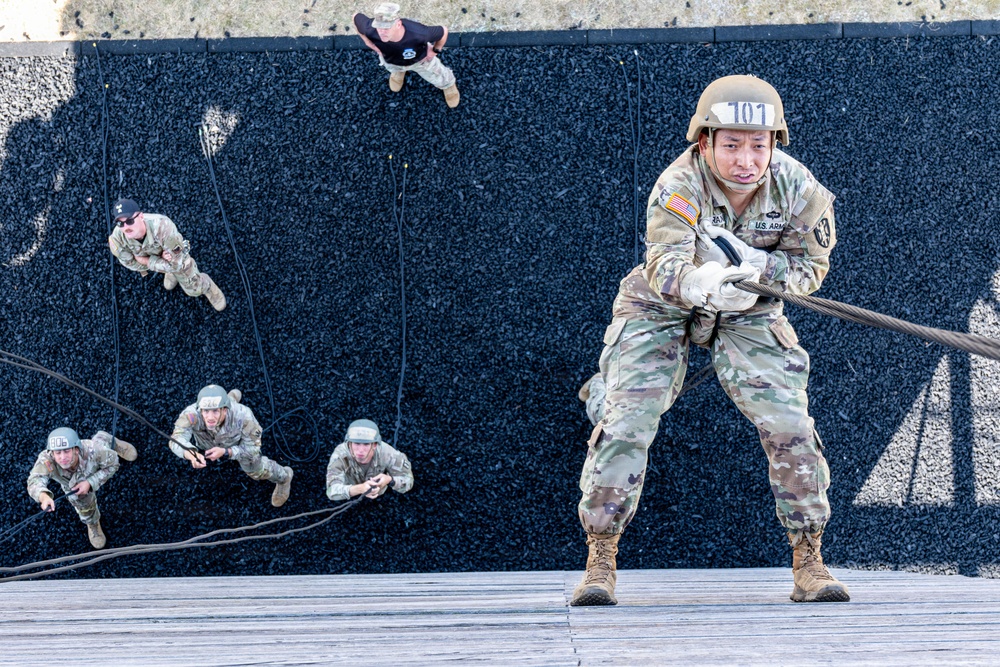 U.S. Army soldiers are conducting a rappelling or rope-climbing training exercise on a large wall covered in black rubber or gravel material. The foreground soldier, numbered '101' and wearing U.S. Army insignia, is at the top of the wall gripping a thick rope, while several other soldiers are visible mid-climb on the wall below. An instructor or observer in a black shirt watches from the top of the wall.