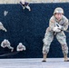U.S. Army soldiers are conducting a rappelling or rope-climbing training exercise on a large wall covered in black rubber or gravel material. The foreground soldier, numbered '101' and wearing U.S. Army insignia, is at the top of the wall gripping a thick rope, while several other soldiers are visible mid-climb on the wall below. An instructor or observer in a black shirt watches from the top of the wall.