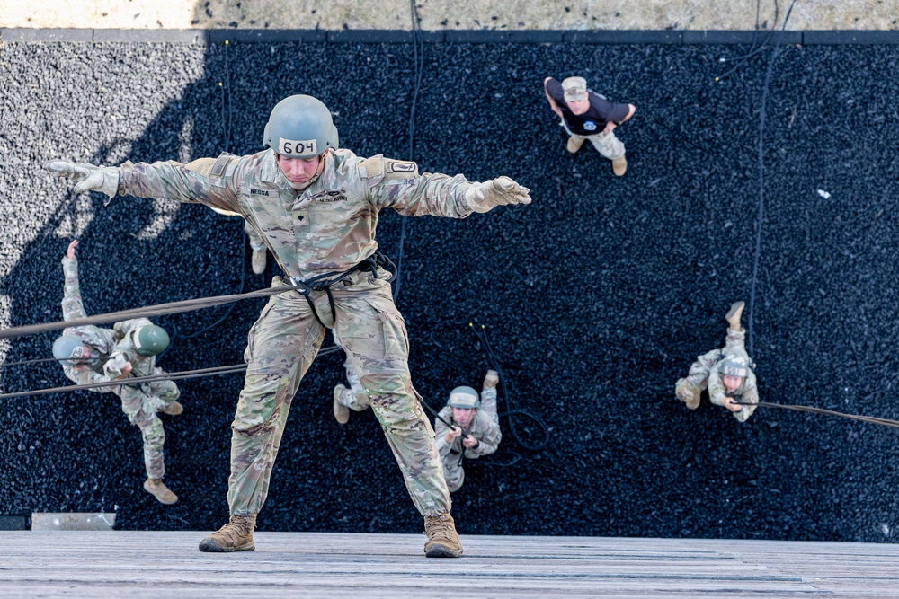 Military personnel are conducting rappelling training on a large wall structure. The central figure, identified by helmet number '604' and name tape 'NESSA', is descending face-forward in a Australian/commando rappel stance with arms outstretched. Multiple other trainees and at least one instructor in civilian attire are visible at various stages of the exercise.