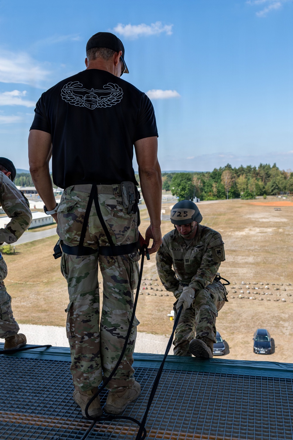 A military training exercise is taking place on top of an elevated platform, likely a rappelling tower. An instructor in a black t-shirt with a harness is supervising a U.S. Army soldier wearing a helmet numbered '725' and identified as 'HEDLICK' who is preparing to rappel. The setting appears to be a military training facility with open fields and tree lines visible in the background.