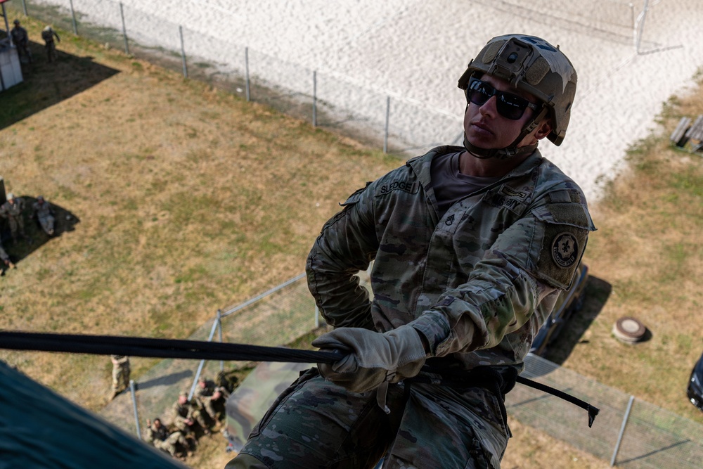 A U.S. Army soldier wearing multicam camouflage, a tactical helmet, and sunglasses is rappelling down a structure during what appears to be a training exercise. The name tape 'SLEDGE' is visible on the soldier's chest. Other soldiers and observers are visible on the ground below.