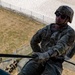 A U.S. Army soldier wearing multicam camouflage, a tactical helmet, and sunglasses is rappelling down a structure during what appears to be a training exercise. The name tape 'SLEDGE' is visible on the soldier's chest. Other soldiers and observers are visible on the ground below.