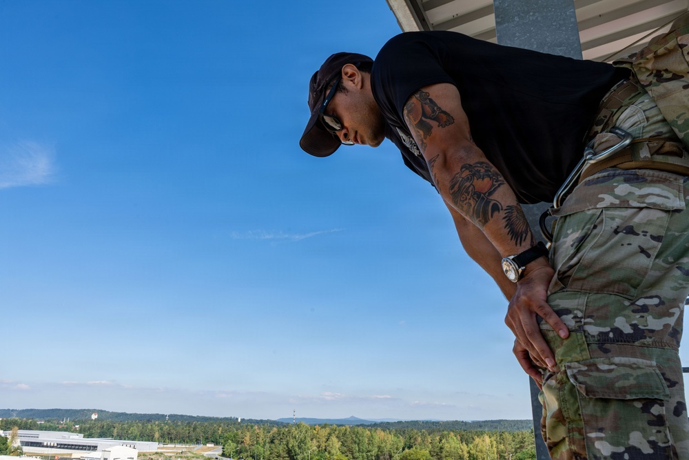 A man wearing a black t-shirt, baseball cap, and sunglasses is leaning over what appears to be a rooftop or elevated platform. He has extensive tattoos on his right arm and is wearing camouflage military-style pants with a watch on his wrist. The background shows a clear blue sky, forested landscape, and distant buildings or structures.
