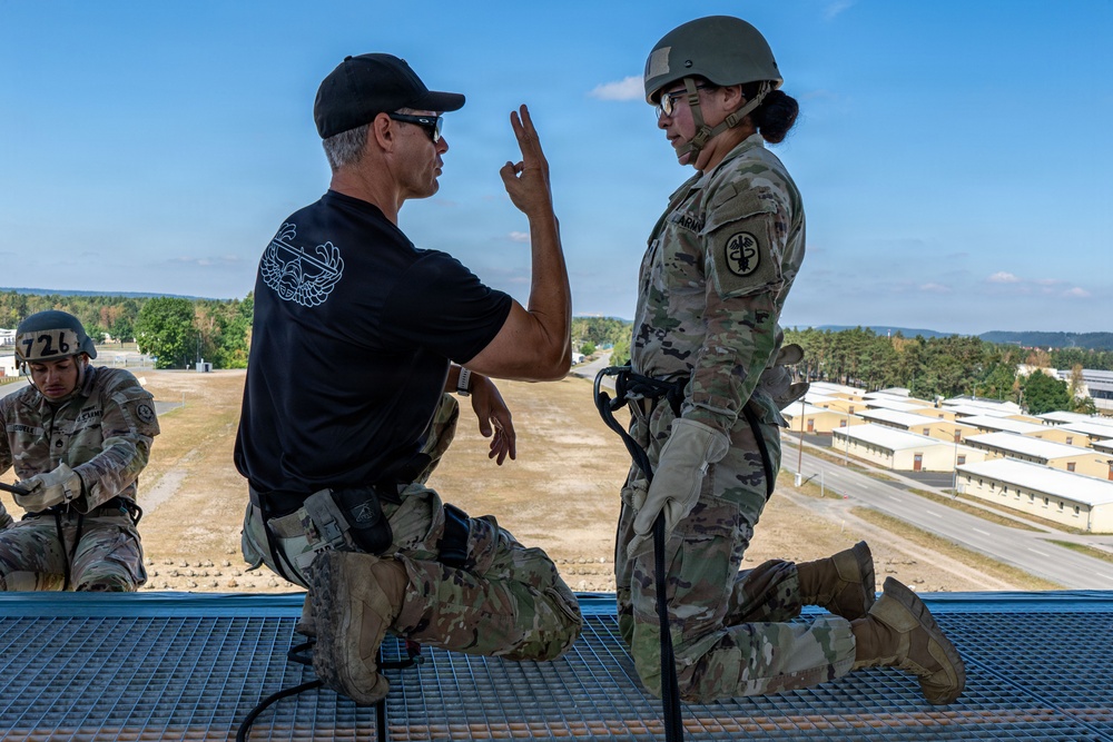 A military instructor in a black t-shirt is briefing or instructing a soldier in full combat gear (OCP uniform, helmet, rappelling harness) on what appears to be the top of a rappelling tower. The instructor is gesturing with raised fingers, likely explaining safety procedures or commands. A second soldier in the background appears to be preparing equipment near the edge of the elevated platform.