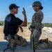 A military instructor in a black t-shirt is briefing or instructing a soldier in full combat gear (OCP uniform, helmet, rappelling harness) on what appears to be the top of a rappelling tower. The instructor is gesturing with raised fingers, likely explaining safety procedures or commands. A second soldier in the background appears to be preparing equipment near the edge of the elevated platform.