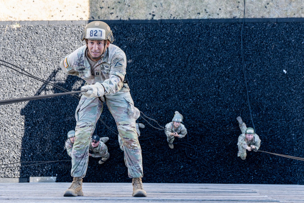 U.S. Army soldiers are conducting a rappelling exercise down a dark textured wall, likely during a military training event or competition. The foreground soldier, numbered '827' with the name 'ZARATE' visible on their uniform, is actively descending while gripping the rope with gloved hands. Three additional soldiers are visible in the background, also rappelling down the same wall.