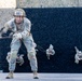 U.S. Army soldiers are conducting a rappelling exercise down a dark textured wall, likely during a military training event or competition. The foreground soldier, numbered '827' with the name 'ZARATE' visible on their uniform, is actively descending while gripping the rope with gloved hands. Three additional soldiers are visible in the background, also rappelling down the same wall.