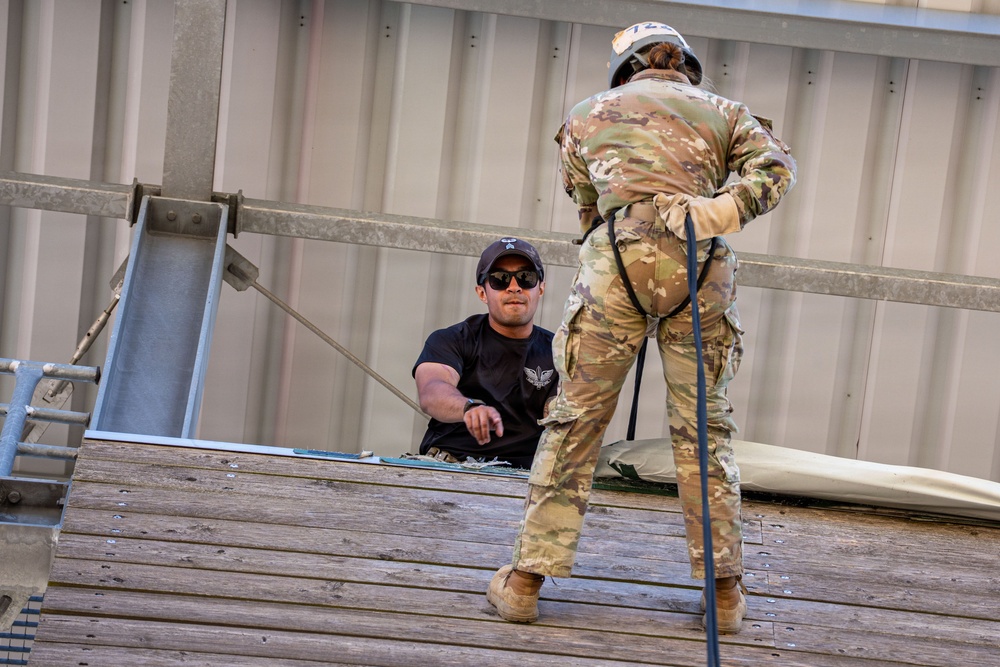 Two individuals are engaged in what appears to be a military rappelling or rope descent training exercise on an elevated wooden platform. One person in OCP (Operational Camouflage Pattern) military uniform and helmet is harnessed with a rope, preparing to rappel, while a second person in a black t-shirt and cap with sunglasses observes and supervises from a crouching position. The setting appears to be a military training tower or facility.