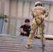 Two individuals are engaged in what appears to be a military rappelling or rope descent training exercise on an elevated wooden platform. One person in OCP (Operational Camouflage Pattern) military uniform and helmet is harnessed with a rope, preparing to rappel, while a second person in a black t-shirt and cap with sunglasses observes and supervises from a crouching position. The setting appears to be a military training tower or facility.
