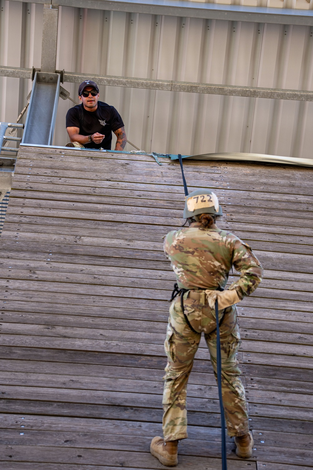 A military training exercise is underway at a rappelling tower. A soldier wearing OCP (Operational Camouflage Pattern) uniform, helmet numbered '722', and a harness is positioned on an inclined wooden wall while holding a rappelling rope. An instructor in a black t-shirt and sunglasses observes from the top of the structure.