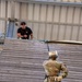 A military training exercise is underway at a rappelling tower. A soldier wearing OCP (Operational Camouflage Pattern) uniform, helmet numbered '722', and a harness is positioned on an inclined wooden wall while holding a rappelling rope. An instructor in a black t-shirt and sunglasses observes from the top of the structure.