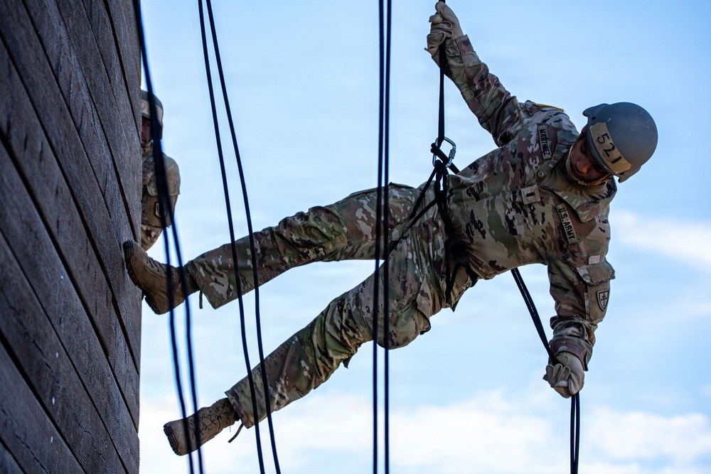 A U.S. Army soldier is rappelling down a tall wooden wall structure during what appears to be a military training exercise. The soldier is wearing full OCP (Operational Camouflage Pattern) uniform with a helmet numbered '521' and is secured by rappelling ropes. A second soldier is partially visible on the left side of the wall, also engaged in the same activity.