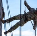 A U.S. Army soldier is rappelling down a tall wooden wall structure during what appears to be a military training exercise. The soldier is wearing full OCP (Operational Camouflage Pattern) uniform with a helmet numbered '521' and is secured by rappelling ropes. A second soldier is partially visible on the left side of the wall, also engaged in the same activity.