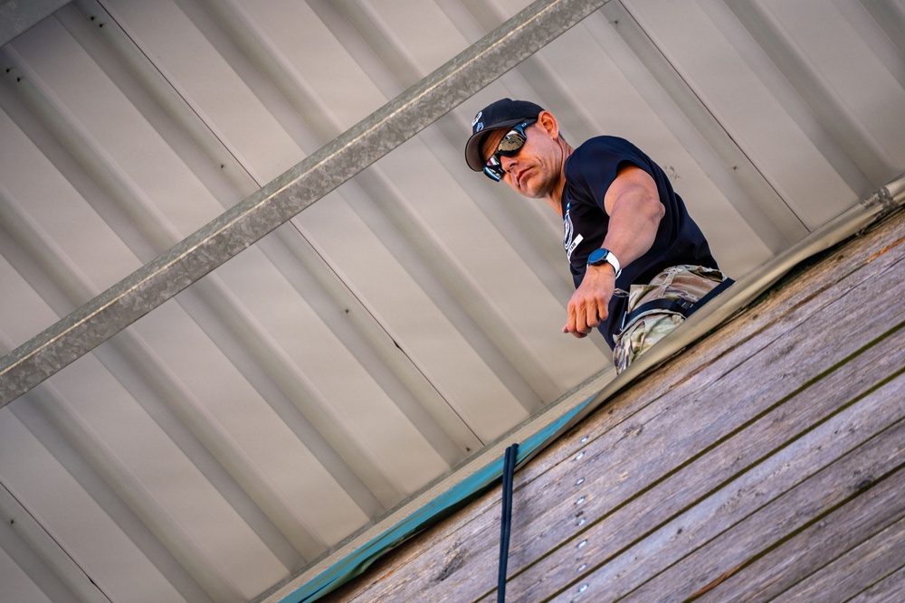 A man is photographed from a low angle while working on or near a rooftop structure. He is wearing a black t-shirt, black baseball cap, reflective sunglasses, camouflage pants, and a blue watch. The perspective suggests the photographer is below, looking up at the subject against a corrugated metal roof or wall.