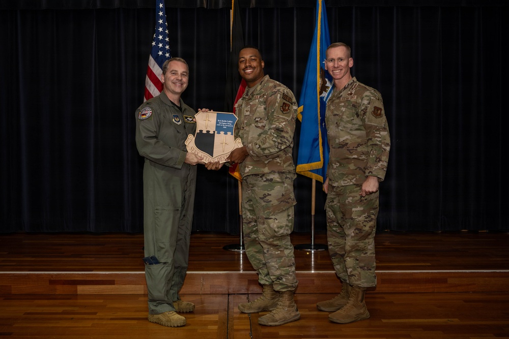 Three U.S. Air Force personnel are posed on a stage during what appears to be an awards or recognition ceremony. The central figure is holding a shield-shaped plaque while flanked by two other service members. An American flag and a blue Air Force flag are visible in the background.