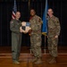 Three U.S. Air Force personnel are posed on a stage during what appears to be an awards or recognition ceremony. The central figure is holding a shield-shaped plaque while flanked by two other service members. An American flag and a blue Air Force flag are visible in the background.