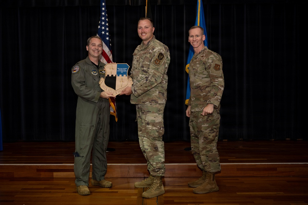 Three military personnel are posed for a formal award or recognition ceremony on a stage. Two individuals in camouflage (OCP) uniforms and one in an olive green flight suit are jointly holding a wooden shield-shaped plaque. An American flag and a blue Air Force flag are visible in the background.