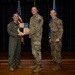 Three military personnel are posed for a formal award or recognition ceremony on a stage. Two individuals in camouflage (OCP) uniforms and one in an olive green flight suit are jointly holding a wooden shield-shaped plaque. An American flag and a blue Air Force flag are visible in the background.