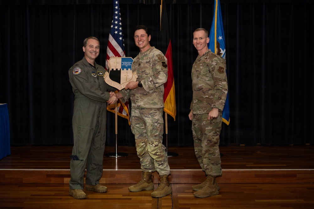 Three military personnel are posed on a stage during what appears to be an awards or recognition ceremony. The central figure is holding a shield-shaped plaque while shaking hands with the person on the left, who is wearing a green flight suit. A US Air Force flag, American flag, and German flag are visible in the background.
