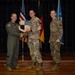 Three military personnel are posed on a stage during what appears to be an awards or recognition ceremony. The central figure is holding a shield-shaped plaque while shaking hands with the person on the left, who is wearing a green flight suit. A US Air Force flag, American flag, and German flag are visible in the background.