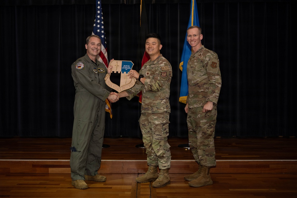 Three military personnel are posed on a stage during what appears to be an awards or recognition ceremony. The central figure is receiving a wooden shield-shaped plaque while shaking hands with an officer in a flight suit on the left. An American flag and a blue military unit flag are visible in the background.