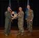 Three military personnel are posed on a stage during what appears to be an awards or recognition ceremony. The central figure is receiving a wooden shield-shaped plaque while shaking hands with an officer in a flight suit on the left. An American flag and a blue military unit flag are visible in the background.