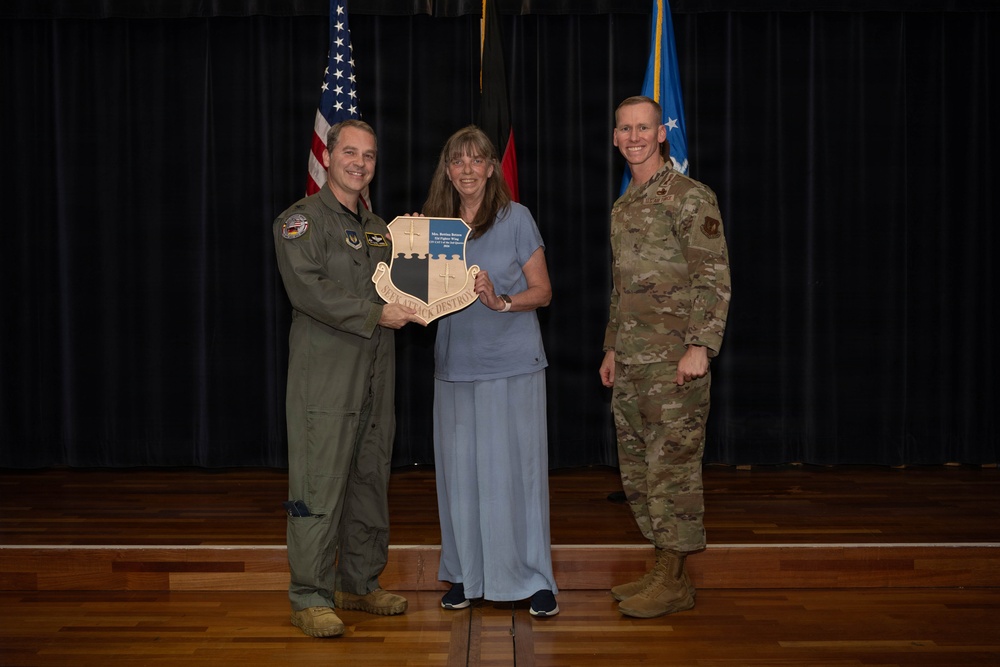 Three individuals pose for a formal photo on a stage, with two military personnel in uniform flanking a civilian woman in the center. The woman is holding a wooden shield-shaped plaque or award, suggesting this is a recognition or award ceremony. The setting appears to be a military installation auditorium or ceremony hall.