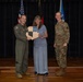 Three individuals pose for a formal photo on a stage, with two military personnel in uniform flanking a civilian woman in the center. The woman is holding a wooden shield-shaped plaque or award, suggesting this is a recognition or award ceremony. The setting appears to be a military installation auditorium or ceremony hall.