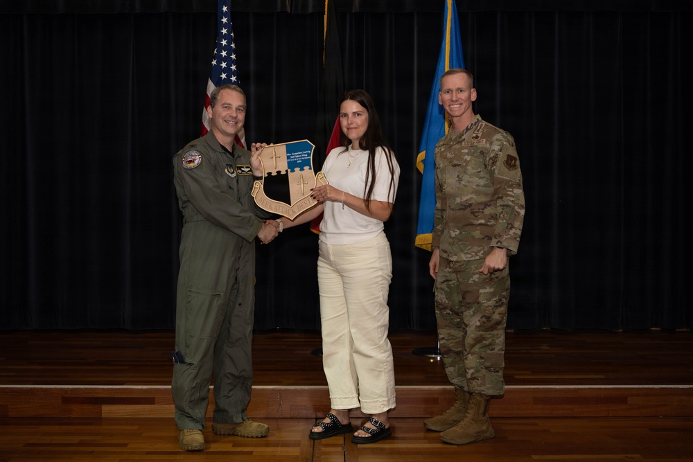 Three individuals pose for a formal photo on a stage during what appears to be a military recognition or award ceremony. A civilian woman in white clothing is receiving a shield-shaped wooden plaque from a military officer in an olive flight suit, while a second officer in camouflage uniform stands to the right. The backdrop includes an American flag and a light blue flag, consistent with a U.S. Air Force setting.