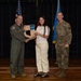 Three individuals pose for a formal photo on a stage during what appears to be a military recognition or award ceremony. A civilian woman in white clothing is receiving a shield-shaped wooden plaque from a military officer in an olive flight suit, while a second officer in camouflage uniform stands to the right. The backdrop includes an American flag and a light blue flag, consistent with a U.S. Air Force setting.