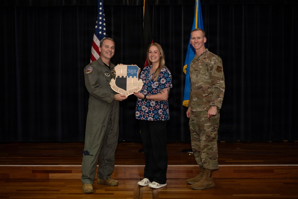 Three individuals pose for a formal photo on a stage, with two military personnel flanking a civilian woman. The group is holding a shield-shaped plaque bearing the motto 'SEEK ATTACK DESTROY,' suggesting an award or recognition ceremony. An American flag and a light blue military flag are visible in the background.