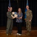 Three individuals pose for a formal photo on a stage, with two military personnel flanking a civilian woman. The group is holding a shield-shaped plaque bearing the motto 'SEEK ATTACK DESTROY,' suggesting an award or recognition ceremony. An American flag and a light blue military flag are visible in the background.