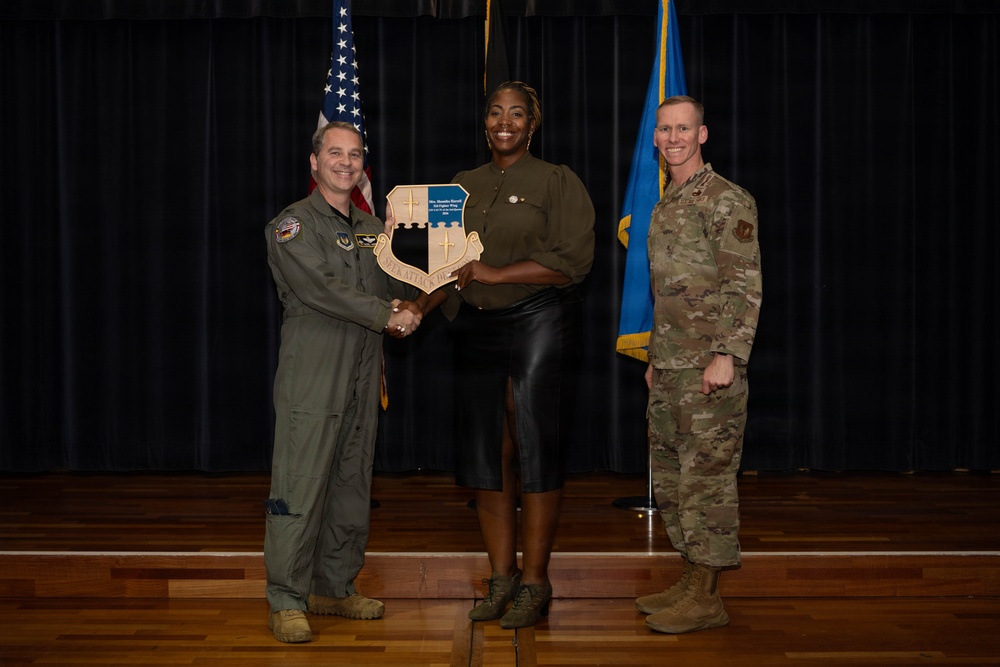 Three individuals are posed on a stage during what appears to be a military award or recognition ceremony. A civilian woman in the center is holding a shield-shaped plaque while shaking hands with a military officer in a flight suit on her left, as another uniformed service member stands to her right. Two flags — an American flag and what appears to be a U.S. Air Force flag — are visible in the background.