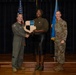Three individuals are posed on a stage during what appears to be a military award or recognition ceremony. A civilian woman in the center is holding a shield-shaped plaque while shaking hands with a military officer in a flight suit on her left, as another uniformed service member stands to her right. Two flags — an American flag and what appears to be a U.S. Air Force flag — are visible in the background.