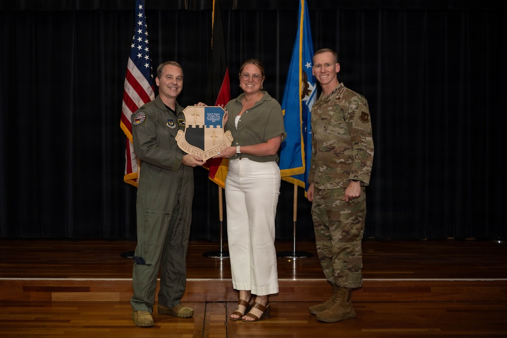 Three individuals pose for a formal photo on a stage, with two military personnel in uniform flanking a civilian woman in the center. They are jointly holding a shield-shaped plaque or award bearing the motto 'SEEK ATTACK DESTROY' and what appears to be a unit crest. The setting includes multiple flags including the U.S. flag, an Air Force flag, and other military unit flags in the background.