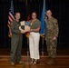 Three individuals pose for a formal photo on a stage, with two military personnel in uniform flanking a civilian woman in the center. They are jointly holding a shield-shaped plaque or award bearing the motto 'SEEK ATTACK DESTROY' and what appears to be a unit crest. The setting includes multiple flags including the U.S. flag, an Air Force flag, and other military unit flags in the background.