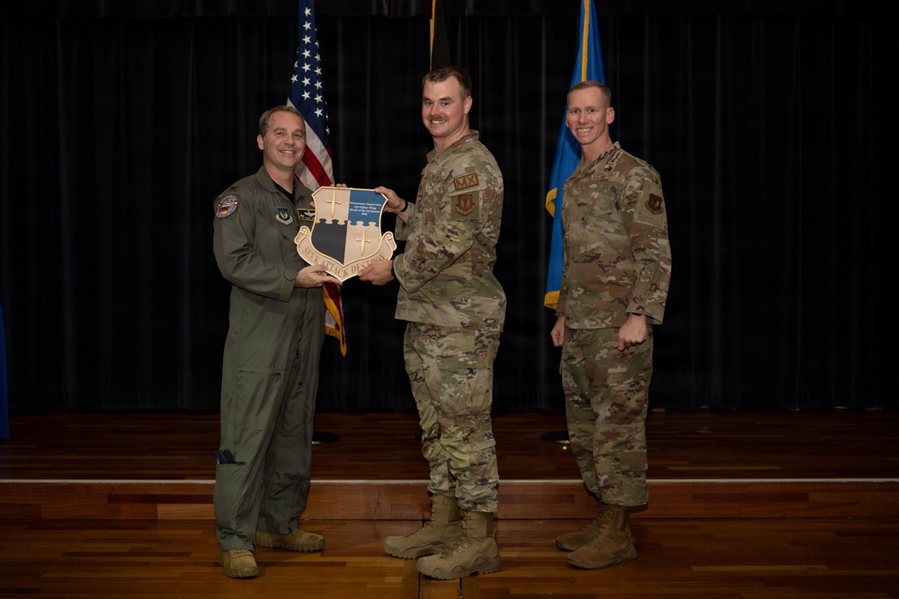 Three military personnel are posed on a stage during what appears to be an awards or recognition ceremony. Two individuals are jointly holding a shield-shaped plaque with the motto 'SEEK ATTACK DESTROY' while a third stands beside them. An American flag and a blue Air Force flag are visible in the background.