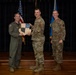 Three military personnel are posed on a stage during what appears to be an awards or recognition ceremony. Two individuals are jointly holding a shield-shaped plaque with the motto 'SEEK ATTACK DESTROY' while a third stands beside them. An American flag and a blue Air Force flag are visible in the background.