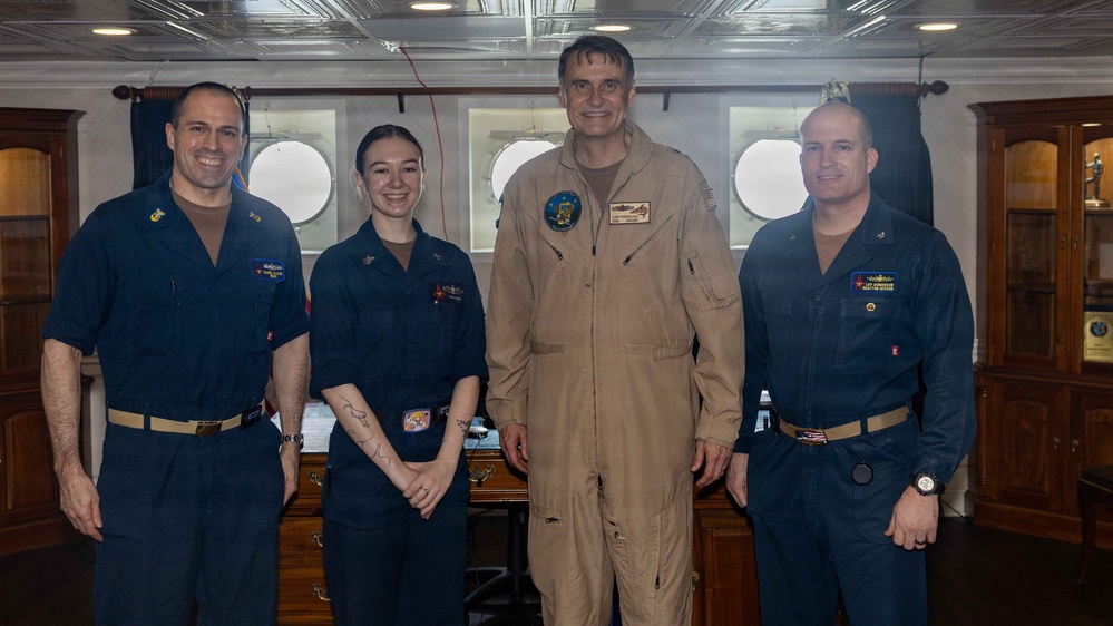Four military personnel pose for a group photo in what appears to be a ship's cabin or officer's quarters. Three individuals wear dark navy blue uniforms while one tall individual in the center wears a tan/khaki flight suit with patches. The setting features porthole windows, wooden cabinetry, and a formal interior consistent with a naval vessel.